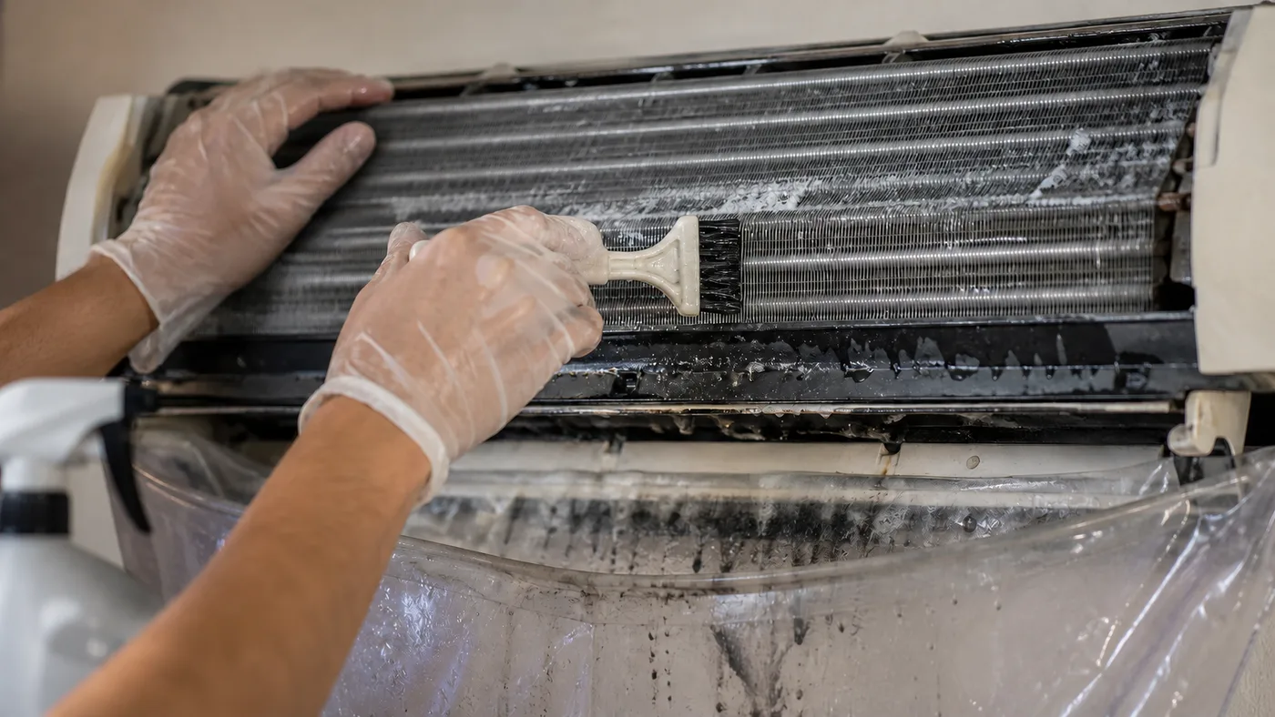Gloved hands brushing the metal fins of an air-conditioner coil, with plastic sheeting below catching the runoff